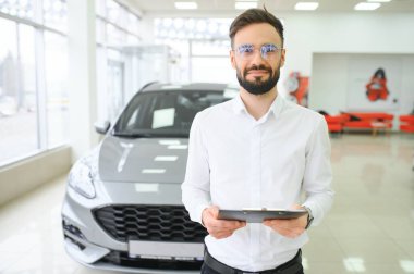 Otomobil galerisindeki resmi kıyafetler içinde elinde pano tutan, kendine güveni tam beyaz bir erkek dükkan asistanının yakın çekimi..