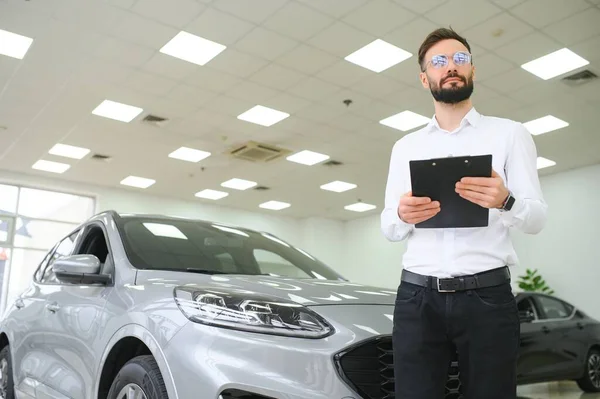 stock image Confident young caucasian businessman shop assistant standing in car automobile dealer shop.