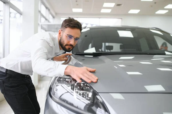 Happy caucasian successful young businessman hugging embracing his new car, feeling excited after buying expensive auto at auto dealer shop store