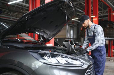 Çalışan üniformalı sakallı bir oto tamircisi bozuk bir arabanın yanında duruyor ve özel bilgisayarı araba bakımı için kullanıyor. Atölyede araba tamir eden bir oto tamircisi