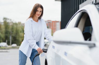 Güneşli alışveriş merkezinin otoparkında elektrik kablosu takan gülümseyen beyaz kadın, kırpıldı. Yaşam tarzı ve ekoloji kavramı