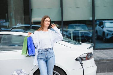 Şehrin otoparkında elektrikli arabanın yanında duran mutlu genç kadın, küçük şehir istasyonundan araba aküsünü şarj ediyor, elinde alışveriş torbaları var.