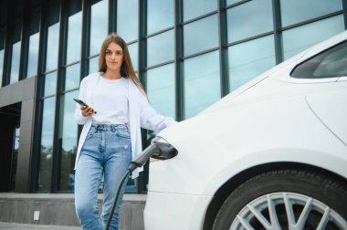 Telefon ellerinde. Elektrikli arabalardaki kadın gündüz şarj istasyonunda. Yepyeni bir araç.