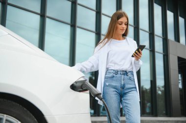 Telefon ellerinde. Elektrikli arabalardaki kadın gündüz şarj istasyonunda. Yepyeni bir araç.