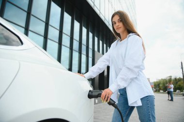 Elinde güç kablosu tutan mutlu genç kız, elektrikli arabanın yanında duruyor, kamu şarj istasyonu.
