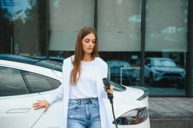 Güneşli alışveriş merkezinin otoparkında elektrik kablosu takan gülümseyen beyaz kadın, kırpıldı. Yaşam tarzı ve ekoloji kavramı