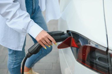 Kocaman gülümseyen genç bir kadın, başka tarafa bakıyor, küçük bir kamu istasyonundan araba aküsünü şarj ediyor, elektrikli arabanın yanında duruyor.