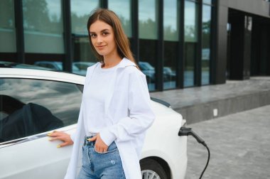 Kocaman gülümseyen genç bir kadın, başka tarafa bakıyor, küçük bir kamu istasyonundan araba aküsünü şarj ediyor, elektrikli arabanın yanında duruyor.
