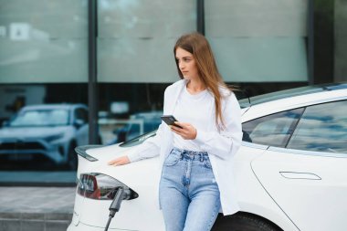 Elektrikli arabanın yanında telefonu olan bir kadın. Araç şarj istasyonunda yüklendi..