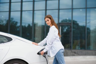 Güneşli alışveriş merkezinin otoparkında elektrik kablosu takan gülümseyen beyaz kadın, kırpıldı. Yaşam tarzı ve ekoloji kavramı