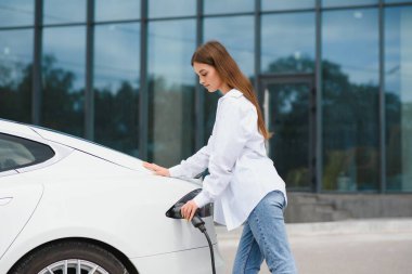 Güneşli alışveriş merkezinin otoparkında elektrik kablosu takan gülümseyen beyaz kadın, kırpıldı. Yaşam tarzı ve ekoloji kavramı
