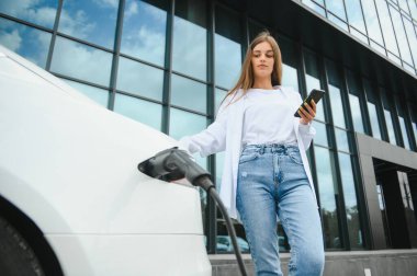 Telefon ellerinde. Elektrikli arabalardaki kadın gündüz şarj istasyonunda. Yepyeni bir araç.