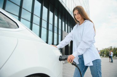 Güneşli alışveriş merkezinin otoparkında elektrik kablosu takan gülümseyen beyaz kadın, kırpıldı. Yaşam tarzı ve ekoloji kavramı