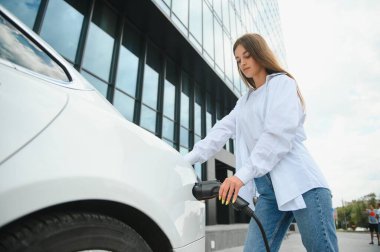 Güneşli alışveriş merkezinin otoparkında elektrik kablosu takan gülümseyen beyaz kadın, kırpıldı. Yaşam tarzı ve ekoloji kavramı