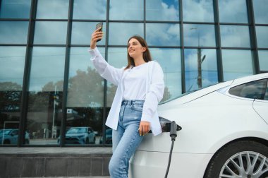 Elektrikli arabanın yanında telefonu olan bir kadın. Araç şarj istasyonunda yüklendi..
