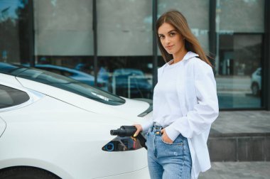 Elinde güç kablosu tutan mutlu genç kız, elektrikli arabanın yanında duruyor, kamu şarj istasyonu.