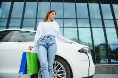 Şehrin otoparkında elektrikli arabanın yanında duran mutlu genç kadın, küçük şehir istasyonundan araba aküsünü şarj ediyor, elinde alışveriş torbaları var.