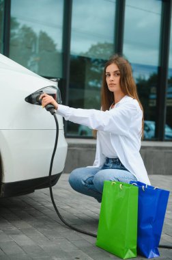 Elinde güç kablosu tutan mutlu genç kız, elektrikli arabanın yanında duruyor, kamu şarj istasyonu.