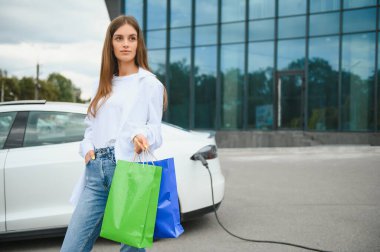 Şehrin otoparkında elektrikli arabanın yanında duran mutlu genç kadın, küçük şehir istasyonundan araba aküsünü şarj ediyor, elinde alışveriş torbaları var.