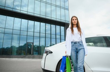 Şehrin otoparkında elektrikli arabanın yanında duran mutlu genç kadın, küçük şehir istasyonundan araba aküsünü şarj ediyor, elinde alışveriş torbaları var.