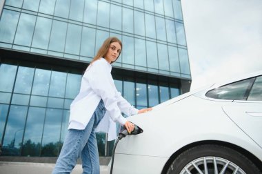 Güneşli alışveriş merkezinin otoparkında elektrik kablosu takan gülümseyen beyaz kadın, kırpıldı. Yaşam tarzı ve ekoloji kavramı