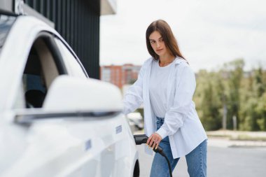 Beyaz bir arabanın arka planında elektrikli bir arabayı şarj etmek için bir kızın eli kablo tutuyor.