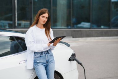 Kocaman gülümseyen genç bir kadın, başka tarafa bakıyor, küçük bir kamu istasyonundan araba aküsünü şarj ediyor, elektrikli arabanın yanında duruyor.