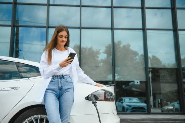 Telefon ellerinde. Elektrikli arabalardaki kadın gündüz şarj istasyonunda. Yepyeni bir araç.