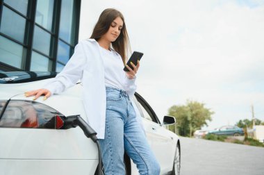 Telefon ellerinde. Elektrikli arabalardaki kadın gündüz şarj istasyonunda. Yepyeni bir araç.
