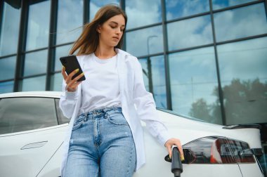 Telefon ellerinde. Elektrikli arabalardaki kadın gündüz şarj istasyonunda. Yepyeni bir araç.
