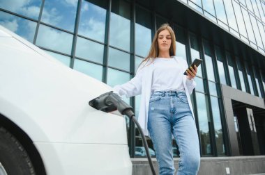 Elektrikli arabanın yanında telefonu olan bir kadın. Araç şarj istasyonunda yüklendi..