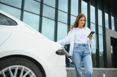 Elektrikli arabanın yanında telefonu olan bir kadın. Araç şarj istasyonunda yüklendi..