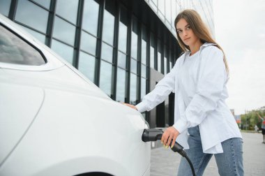 Kocaman gülümseyen genç bir kadın, başka tarafa bakıyor, küçük bir kamu istasyonundan araba aküsünü şarj ediyor, elektrikli arabanın yanında duruyor.