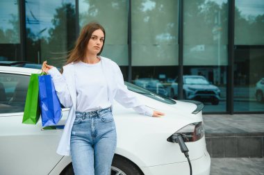 Şehrin otoparkında elektrikli arabanın yanında duran mutlu genç kadın, küçük şehir istasyonundan araba aküsünü şarj ediyor, elinde alışveriş torbaları var.