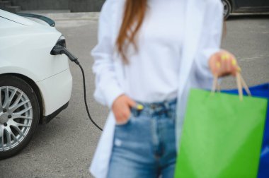 Kocaman gülümseyen genç bir kadın, başka tarafa bakıyor, küçük bir kamu istasyonundan araba aküsünü şarj ediyor, elektrikli arabanın yanında duruyor.