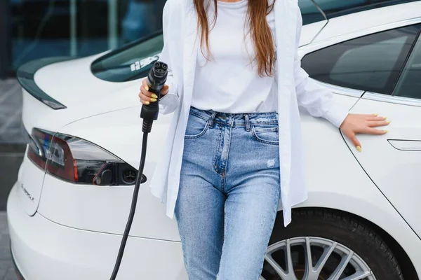 Happy young adult girl holding power cable supply in hand, standing near electric car, public charging station