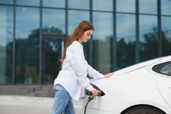 stock image Happy young adult girl holding power cable supply in hand, standing near electric car, public charging station