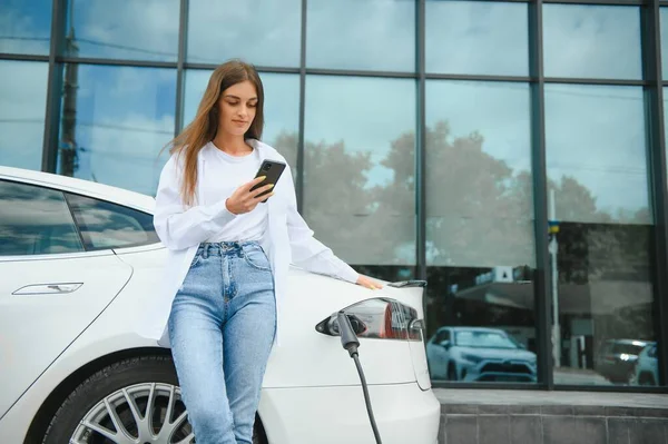 stock image Phone in hands. Woman on the electric cars charge station at daytime. Brand new vehicle