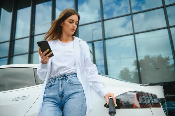 stock image Woman with phone near an rental electric car. Vehicle charged at the charging station.