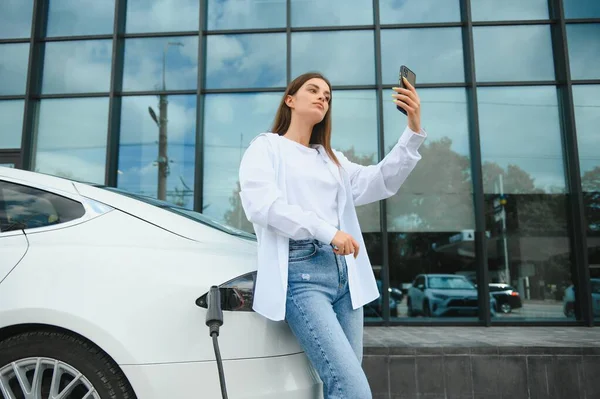 stock image Phone in hands. Woman on the electric cars charge station at daytime. Brand new vehicle