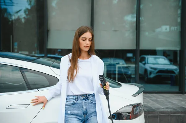 stock image Smiling young caucasian woman plugging electricity cable in electric vehicle for charging on sunny mall parking, cropped. Lifestyle and ecology concept