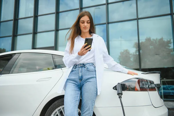 stock image Happy young adult woman smiling wide, looking away, charging automobile battery from small public station, standing near electric car