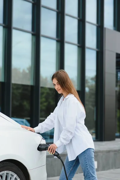 stock image Happy young adult girl holding power cable supply in hand, standing near electric car, public charging station