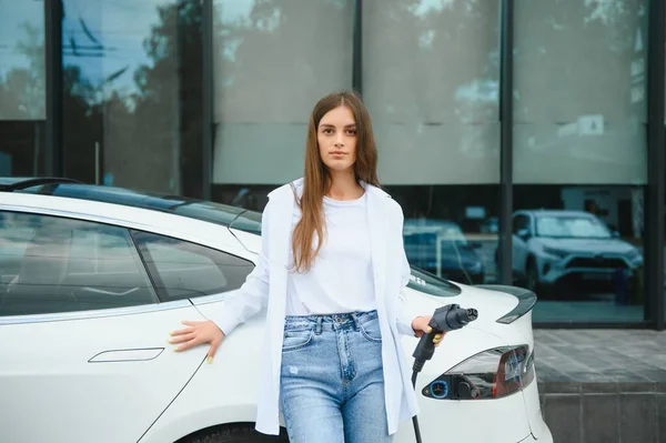 stock image Happy young adult girl holding power cable supply in hand, standing near electric car, public charging station