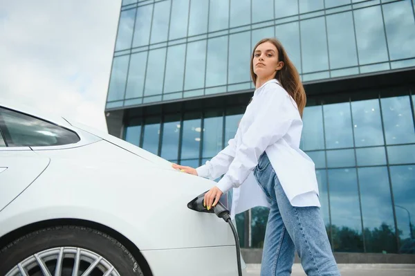 Güneşli alışveriş merkezinin otoparkında elektrik kablosu takan gülümseyen beyaz kadın, kırpıldı. Yaşam tarzı ve ekoloji kavramı