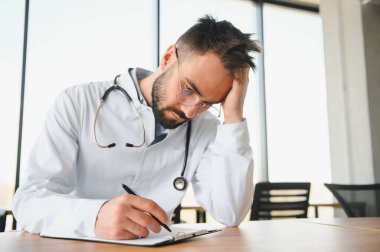 Tıp üniforması giyen yakışıklı doktor iş yerinde oturmaktan yorgun düşmüş başını tutmaktan yorgun düşmüş ve başı ağrıyormuş. Stres ve hüsran kavramı.