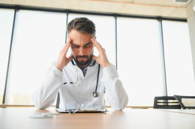 Tıp üniforması giyen yakışıklı doktor iş yerinde oturmaktan yorgun düşmüş başını tutmaktan yorgun düşmüş ve başı ağrıyormuş. Stres ve hüsran kavramı.