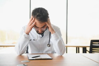 Handsome doctor man wearing medical uniform sitting on his workplace tired holding his head feeling fatigue and headache. Stress and frustration concept.