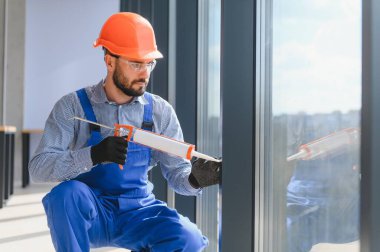 Workers with tube of sealant and suction lifters installing plastic windows indoors