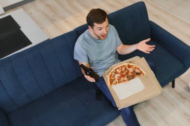 Mutlu genç adam evde TV izlerken bira içip pizza yiyor..